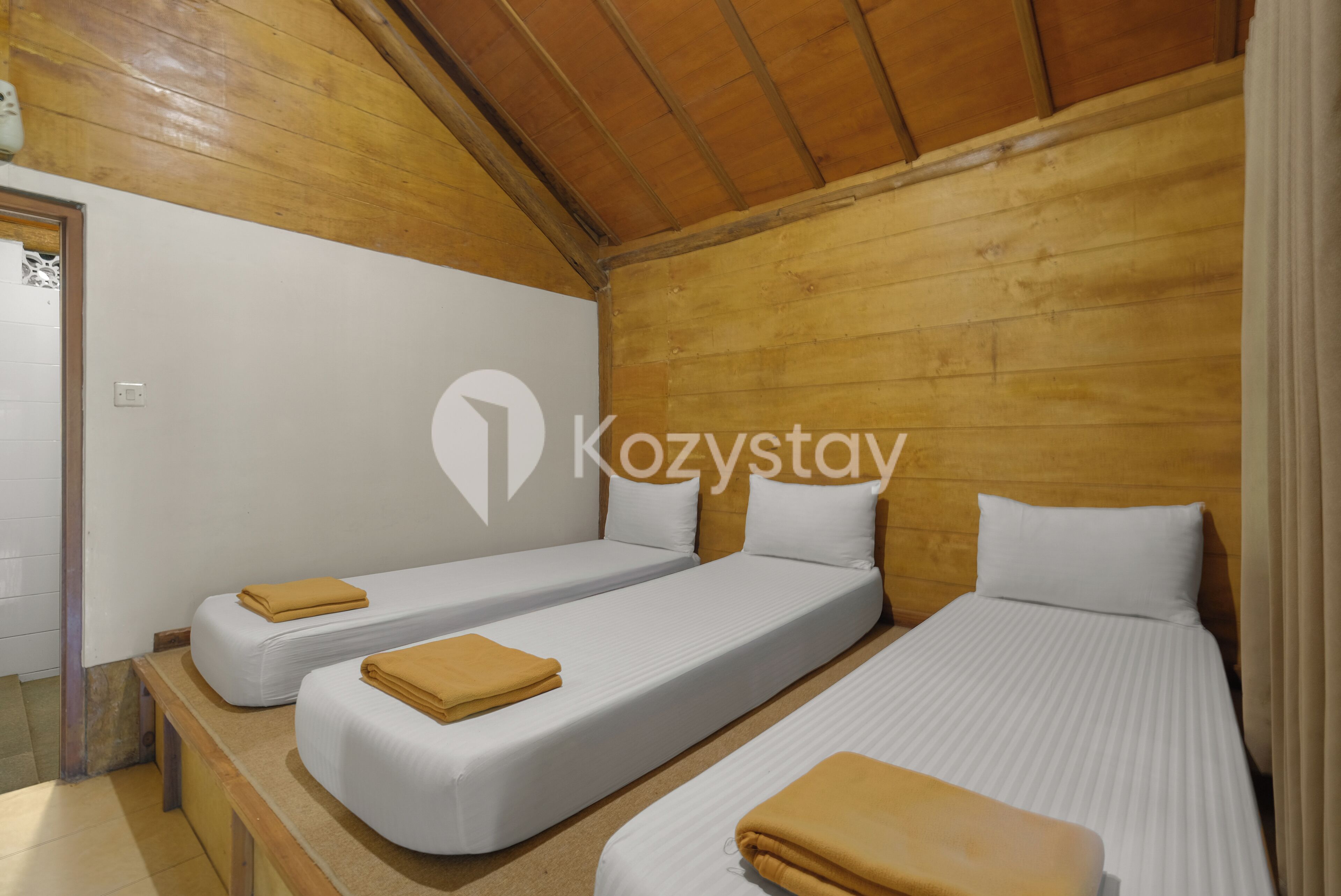 Una habitación con tres camas individuales, cada una con un colchón blanco y una colcha amarilla. La habitación tiene paredes de madera y un techo inclinado.