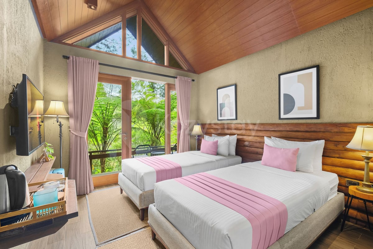 A room with two beds, a wooden ceiling, a large window with curtains, a bedside table with a lamp, and a wall-mounted TV.