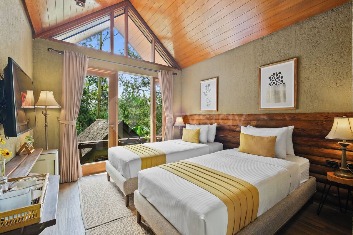 A hotel room with two beds, a desk, a lamp, and a large window with curtains.