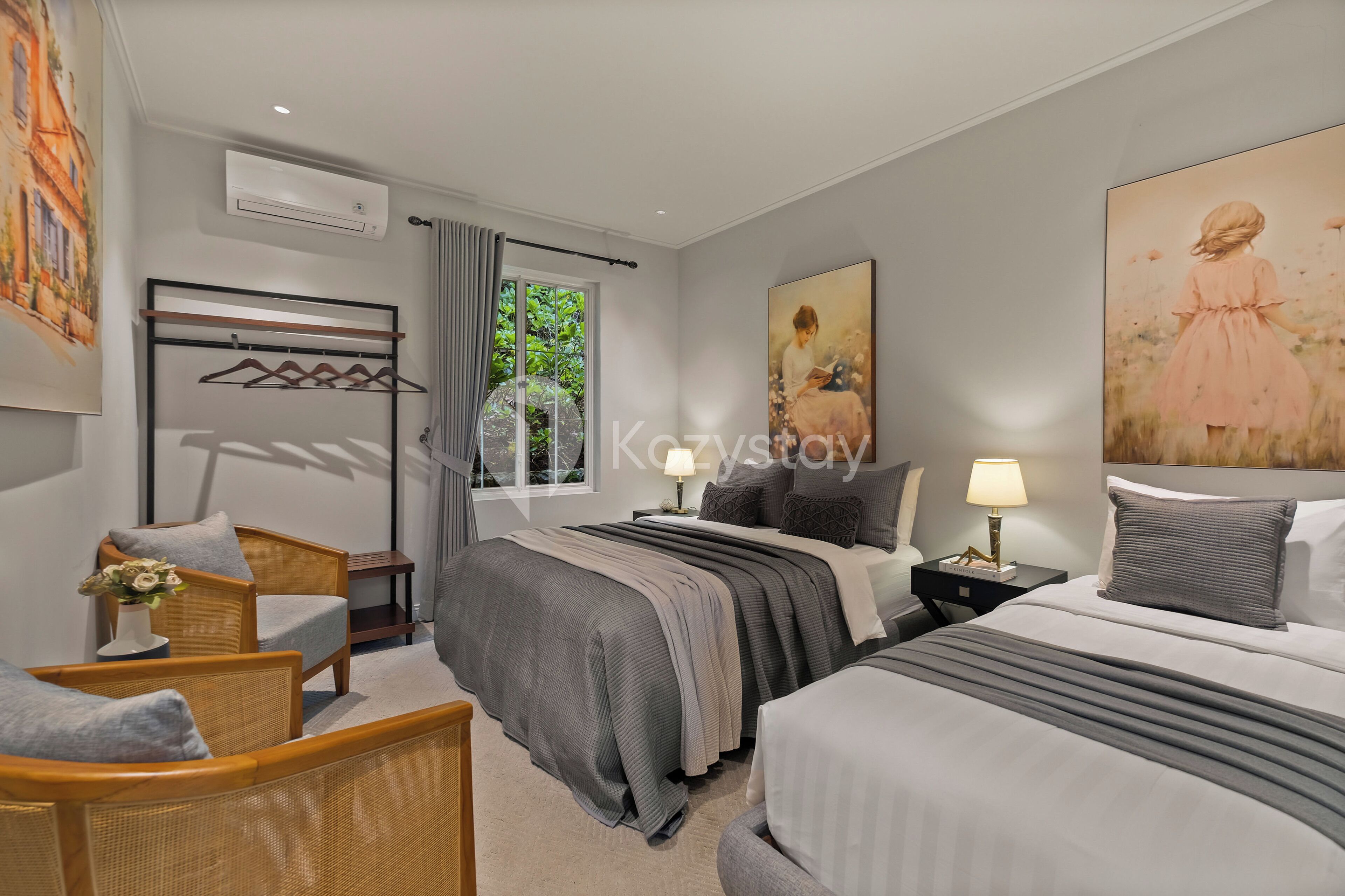 Habitación de hotel con cama, dos mesitas de noche, una silla, ventana con cortinas y obras de arte en las paredes.