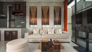 A modern living room with a sofa, coffee table, and a TV. There are decorative wall panels and a kitchen area with cabinets and a tiled backsplash. - PoolVilla Jacuzzi & Mountain View V232 (Rawai)