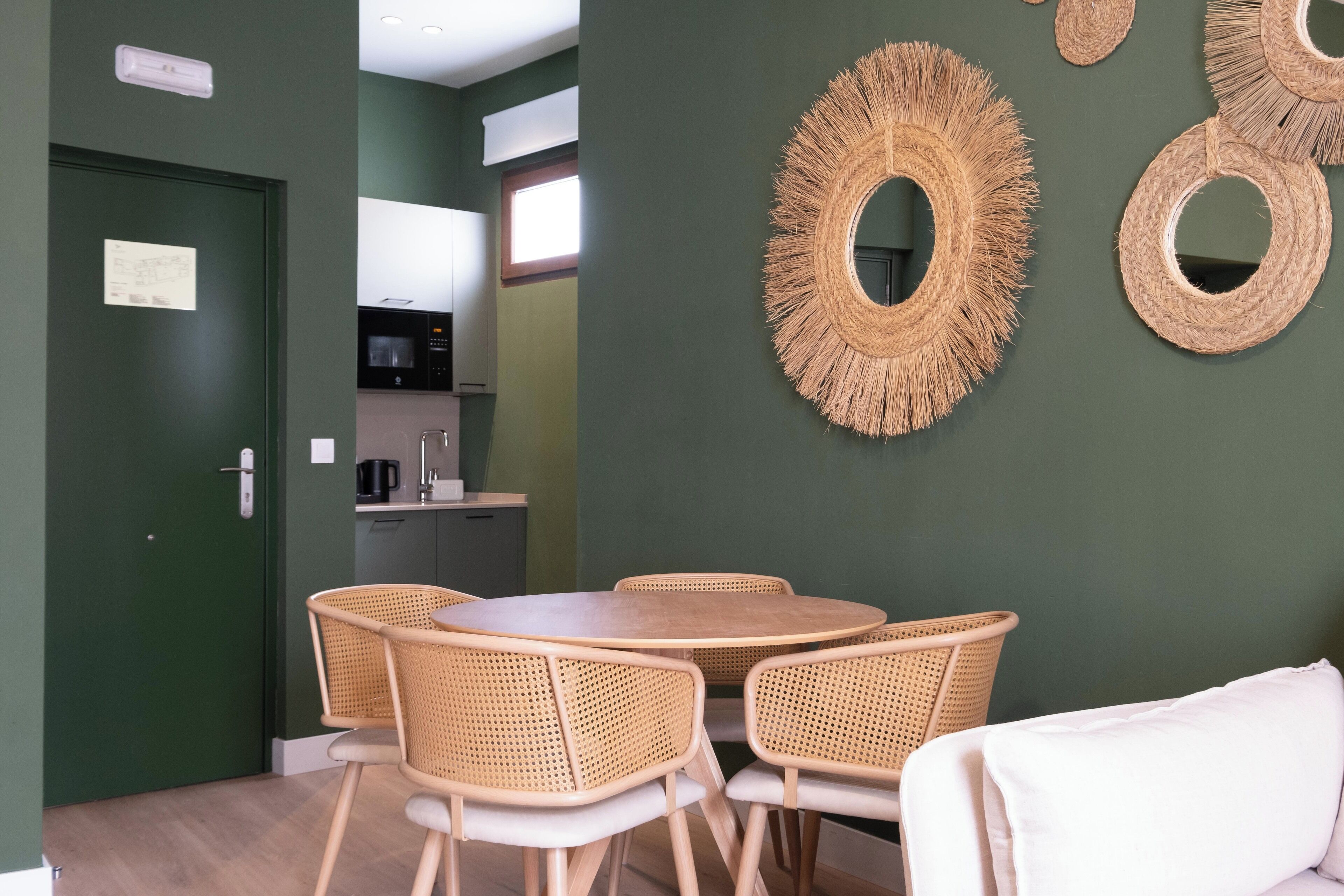 Comedor con una mesa redonda y cuatro sillas de mimbre, una pared verde y una cocina al fondo.