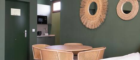 Comedor con una mesa redonda y cuatro sillas de mimbre, una pared verde y una cocina al fondo.
