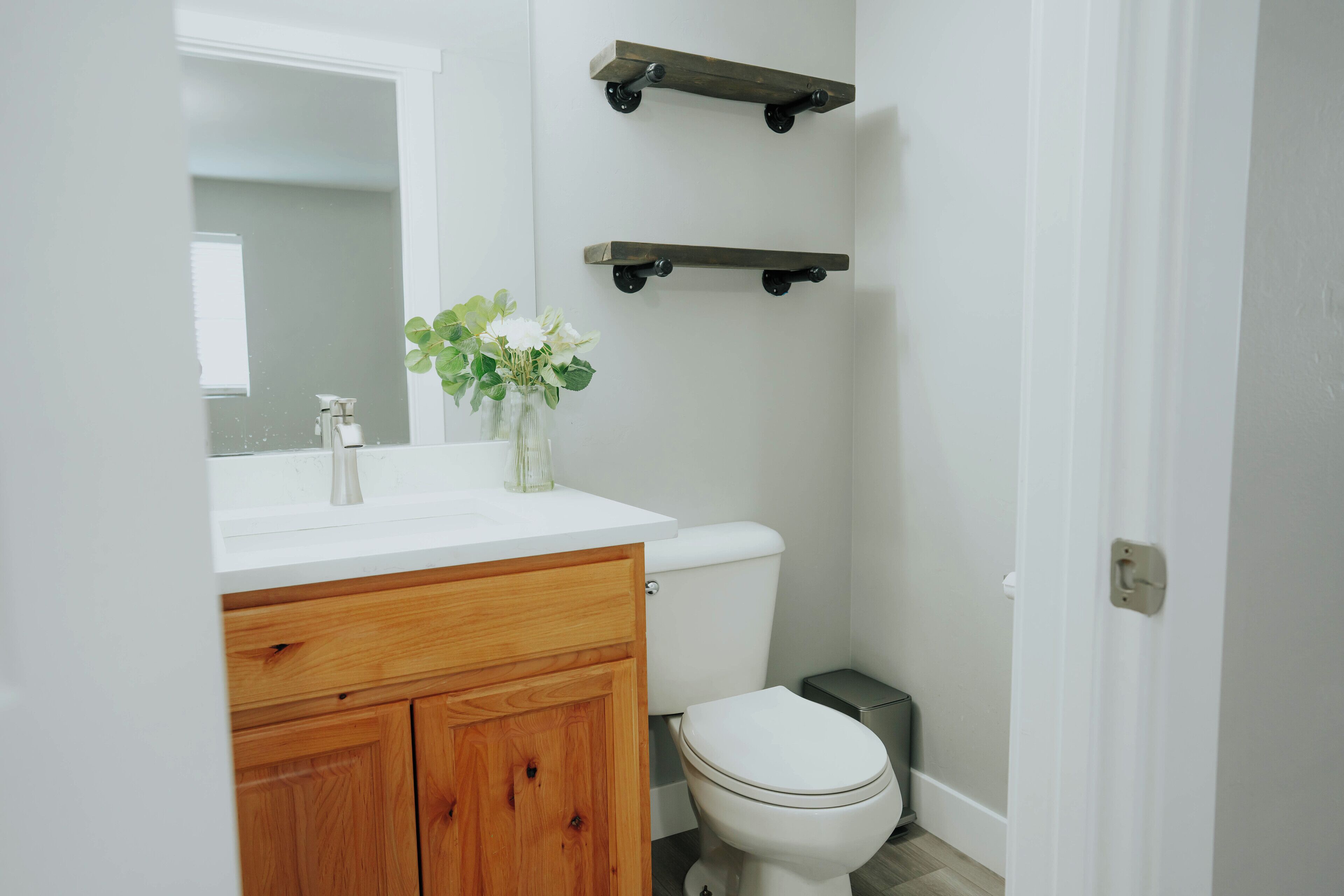 Un baño con inodoro blanco, tocador de madera, lavamanos blanco y espejo. Hay dos estantes de madera montados en la pared sobre el inodoro.