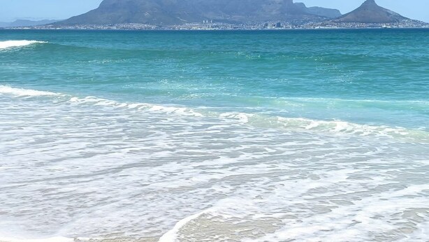 Une plage bordée d’une eau turquoise et transparente, avec une chaîne de montagnes qui se dessine à l’horizon.