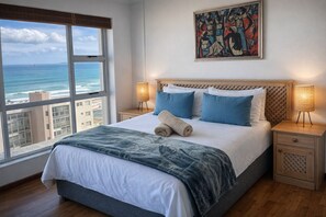 A bedroom with a large window overlooking the ocean, a bed with blue and white bedding, and a painting on the wall. - Views, Views and more Views! (Cape Town)