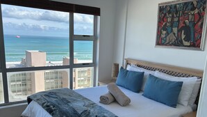 Une chambre d’hôtel dotée d’une grande fenêtre donnant sur l’océan et le paysage urbain. La chambre comprend un lit recouvert d’une courtepointe à motifs foncés, des oreillers bleus et blancs, et un lit parfaitement fait avec des serviettes.