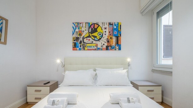 Een hotelkamer met een groot bed, twee nachtkastjes, een schilderij aan de muur en een raam met jaloezieën.