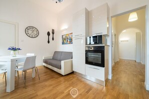 Una cocina moderna con armarios blancos, un microondas y un horno. Hay una zona de comedor con una mesa y sillas blancas, y un sofá gris. Un reloj y una cuchara están colgados en la pared.