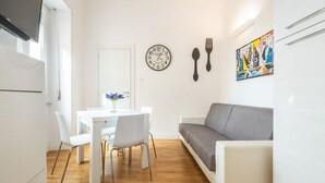 Een knus woongedeelte met een eettafel, stoelen, een grijze bank, een wandklok en een kleurrijk abstract schilderij.