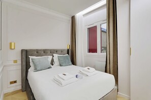 Un lit bien fait, avec du linge de lit et des serviettes blanches, une tête de lit capitonnée et une fenêtre avec des rideaux.