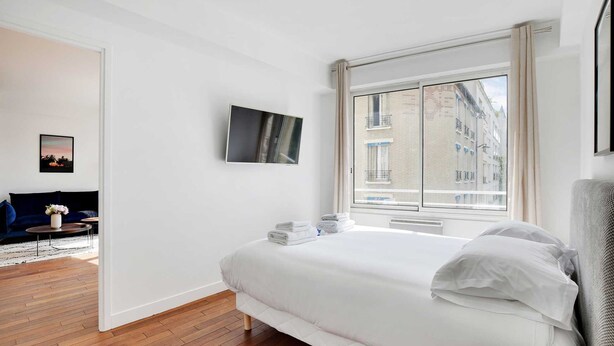 Une chambre moderne avec un grand lit, un téléviseur à écran plat, une vue sur les immeubles par la fenêtre et un canapé de couleur sombre dans la pièce adjacente.