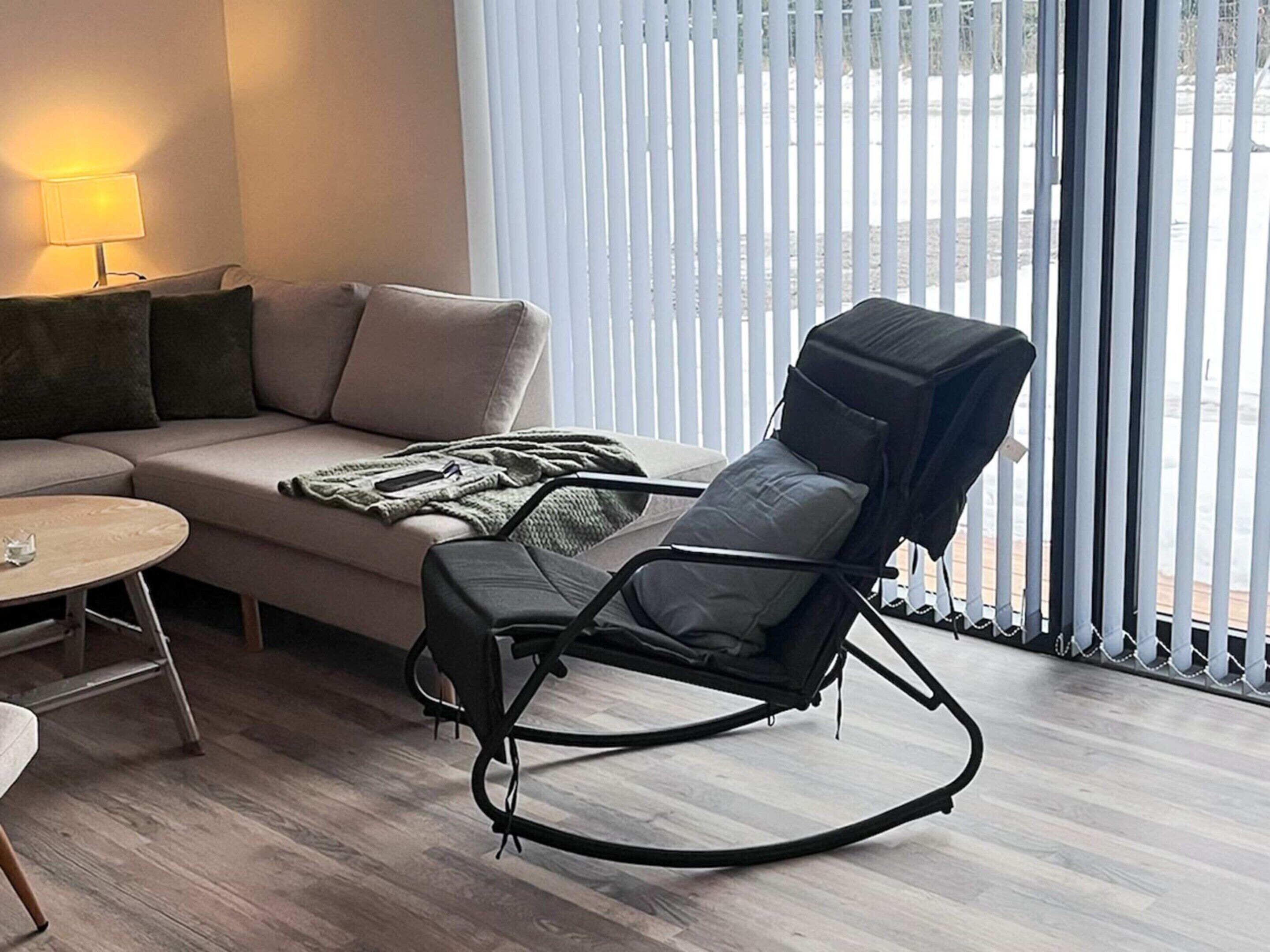 A modern living room with a sofa, a small round table, a black rocking chair, and a window with blinds.