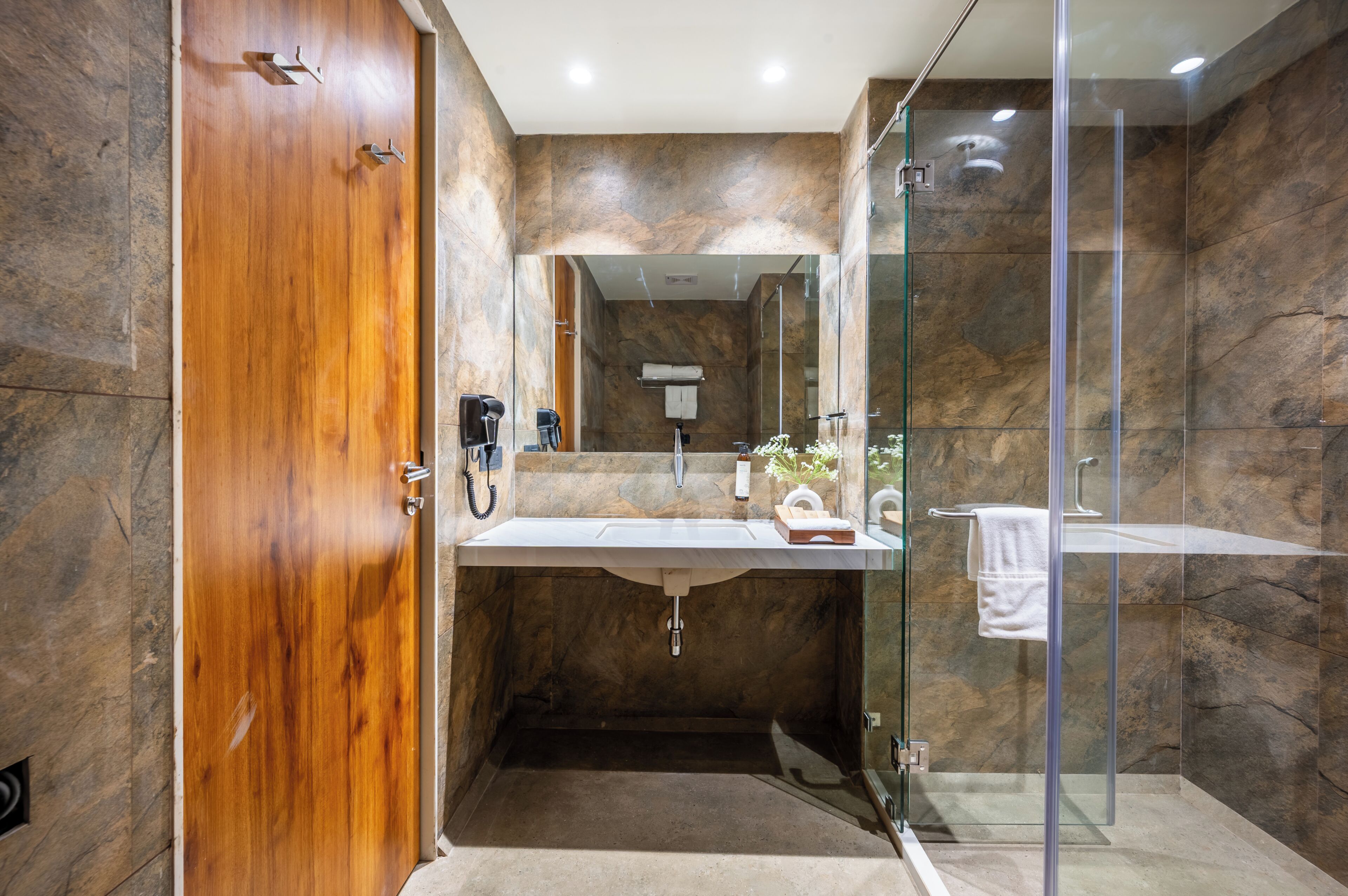 A modern bathroom with a wooden door, a glass shower enclosure, a marble-tiled wall, and a white sink.