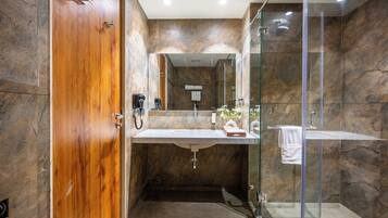 A modern bathroom with a wooden door, a glass shower enclosure, a marble-tiled wall, and a white sink.