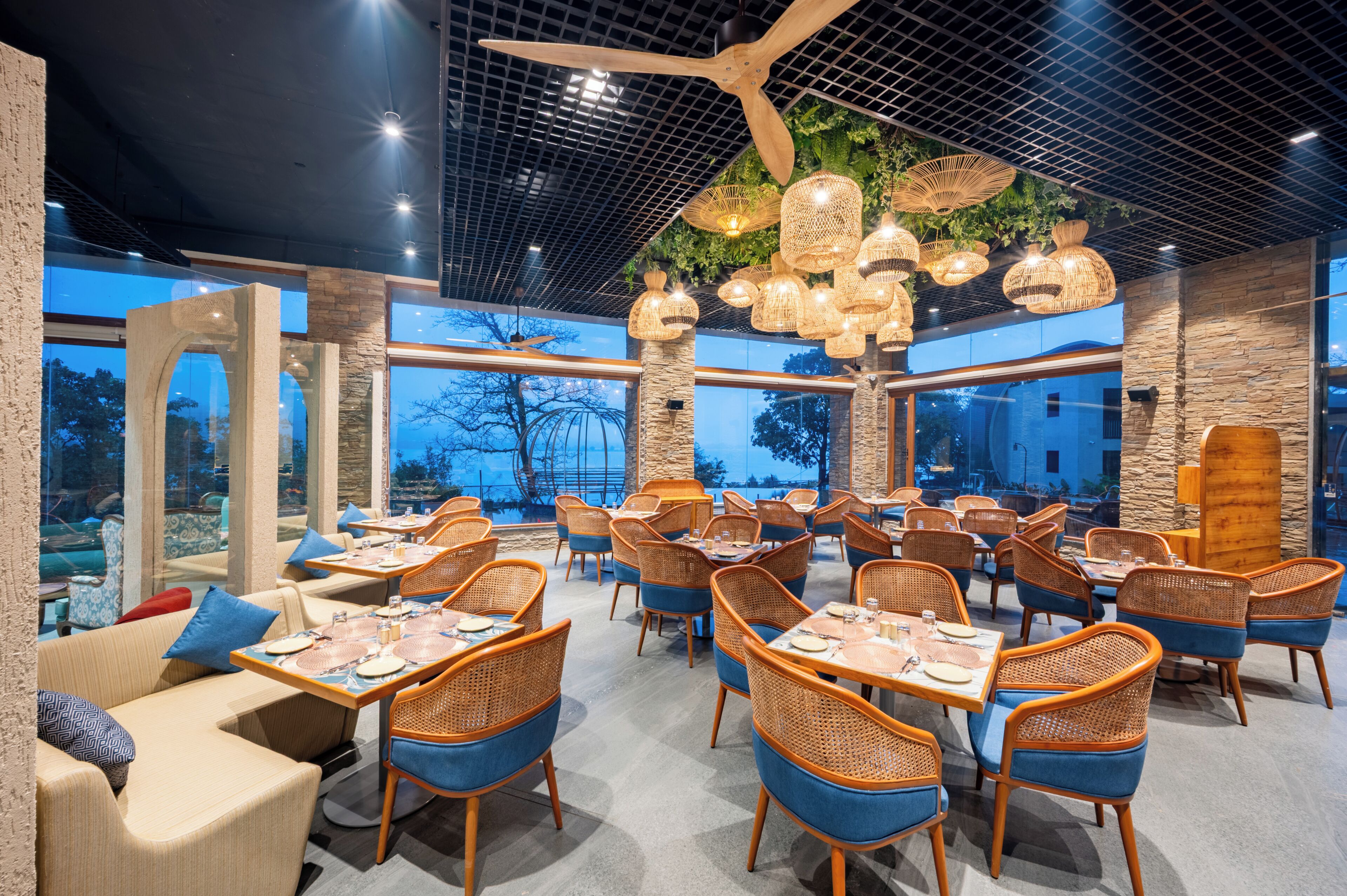 Un restaurante con mobiliario de mimbre, ventanas amplias y lámparas colgantes.