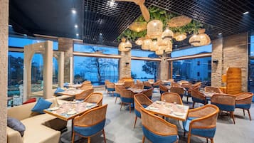Un restaurante con mobiliario de mimbre, ventanas amplias y lámparas colgantes.