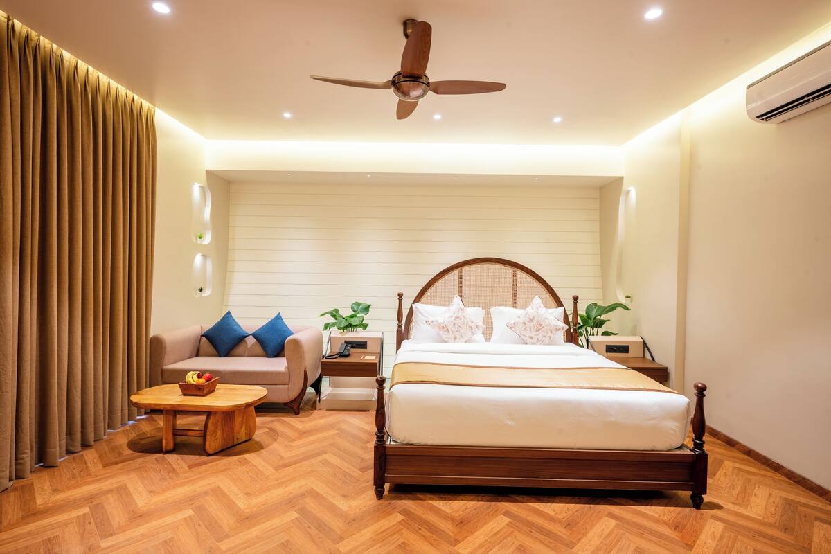 Une chambre à coucher avec un grand lit, une tête de lit en bois, un ventilateur de plafond, une table de chevet avec une lampe, une petite table avec des fruits et un canapé avec des coussins.