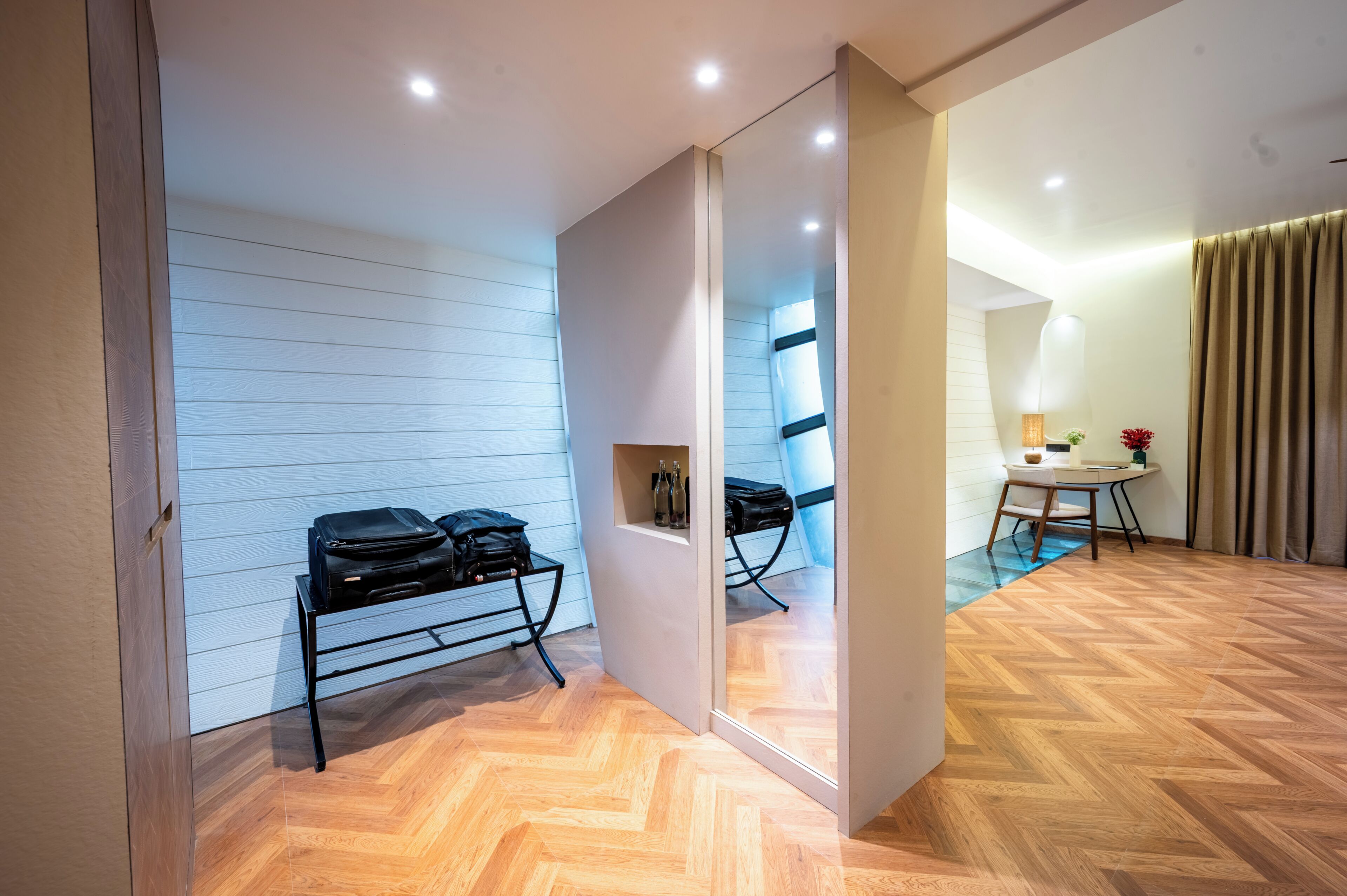 Habitación de hotel con piso de madera, un escritorio con silla, un espejo y dos maletas negras sobre un carrito.