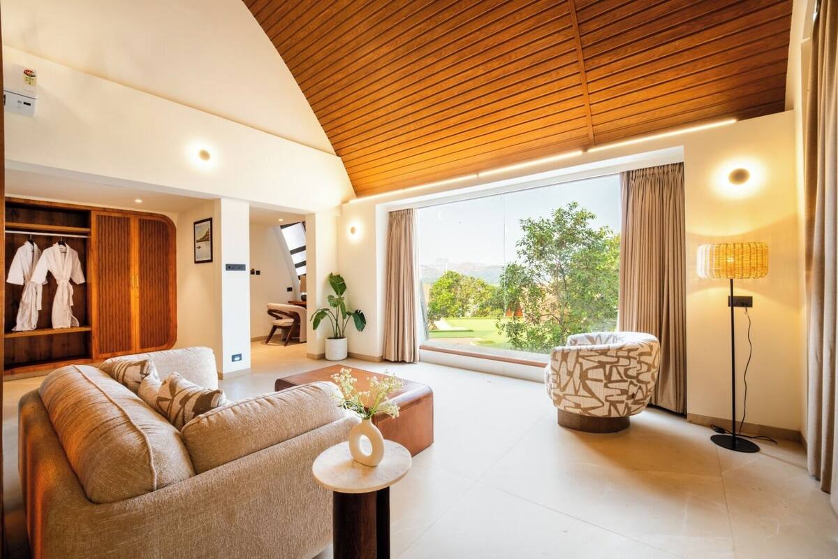 Un vaste espace de vie doté d’un plafond en bois, d’un canapé, d’une table basse, d’un pouf à motifs, d’un lampadaire, d’une plante en pot et offrant une vue sur la verdure grâce à de grandes fenêtres.