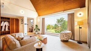 A spacious living area with a wooden ceiling, a sofa, a coffee table, a patterned ottoman, a floor lamp, a potted plant, and a view of greenery through large windows. - ARK Lake Resort (Igatpuri)