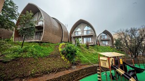 A modern building with unique curved architecture, a playground with slides, and a green artificial pool. - ARK Lake Resort (Igatpuri)