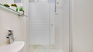 Un bagno stretto con un lavandino bianco, uno scaffale in vetro con piante e un angolo doccia.