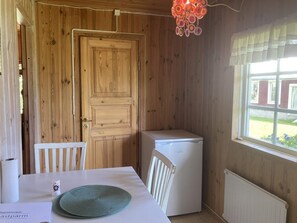 Une pièce aux murs lambrissés, avec une table blanche, une chaise blanche, un réfrigérateur blanc et une fenêtre donnant sur un paysage verdoyant.