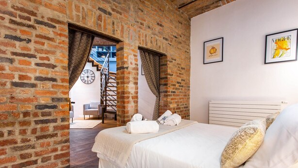 Un dormitorio con pared de ladrillo, una cama con ropa blanca, una escalera y dos cuadros en la pared.