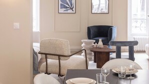 Un comedor con una mesa de madera oscura dispuesta para cuatro personas, rodeada de sillas blancas tapizadas. Se observan obras de arte enmarcadas en las paredes, un sillón azul y un sofá blanco.