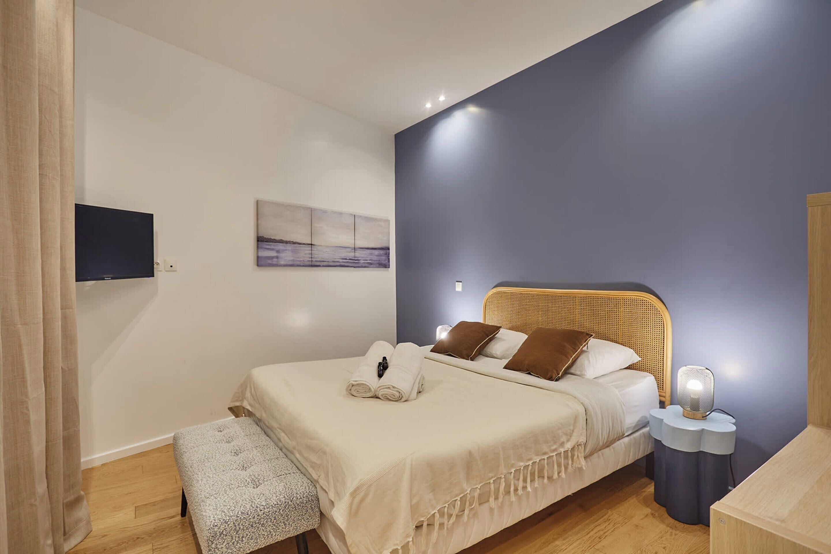 Une chambre à coucher comprenant un lit, une table de chevet, un banc et un téléviseur fixé au mur.
