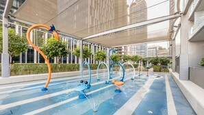 An outdoor water feature with multiple fountains, surrounded by a modern urban setting with buildings and greenery.