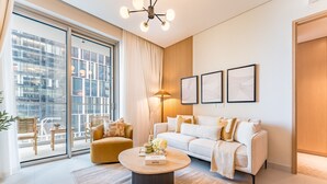 A modern living room with a sofa, armchair, and coffee table. There is a large window with a view of buildings, a chandelier, and framed artwork on the wall.