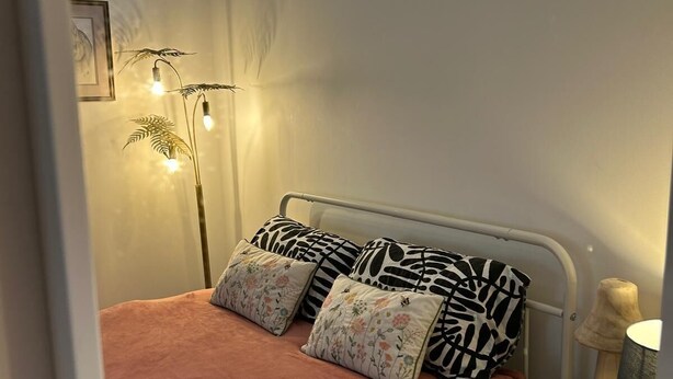 A bedroom with a bed, a nightstand, a lamp, a window with a dog statue, and a plant.