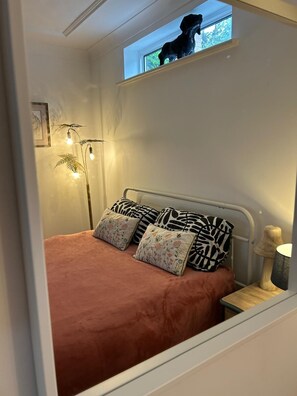 Un dormitorio con cama, mesita de noche, lámpara, ventana y una planta, además de una estatua de perro.