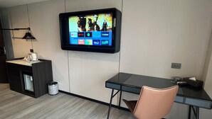 Et moderne hotelværelse med et fladskærms-tv på væggen, der viser forskellige streamingtjenester, et lille tekøkken, et skrivebord med stol og en skraldespand.
