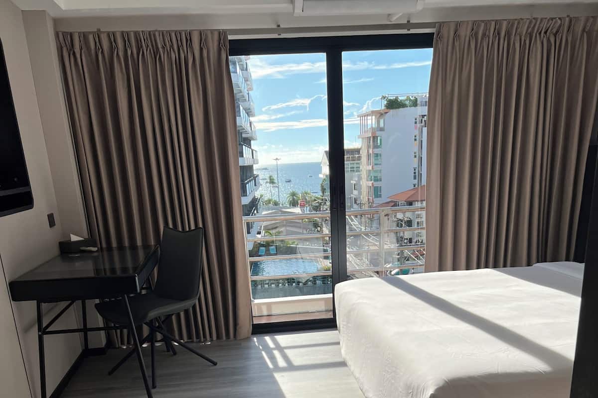Habitación de hotel con cama, escritorio y silla, y vista a edificios y un cuerpo de agua a través de un ventanal grande.