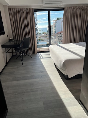 A hotel room with a bed, a desk with a chair, and a view of buildings and a body of water through a large window.