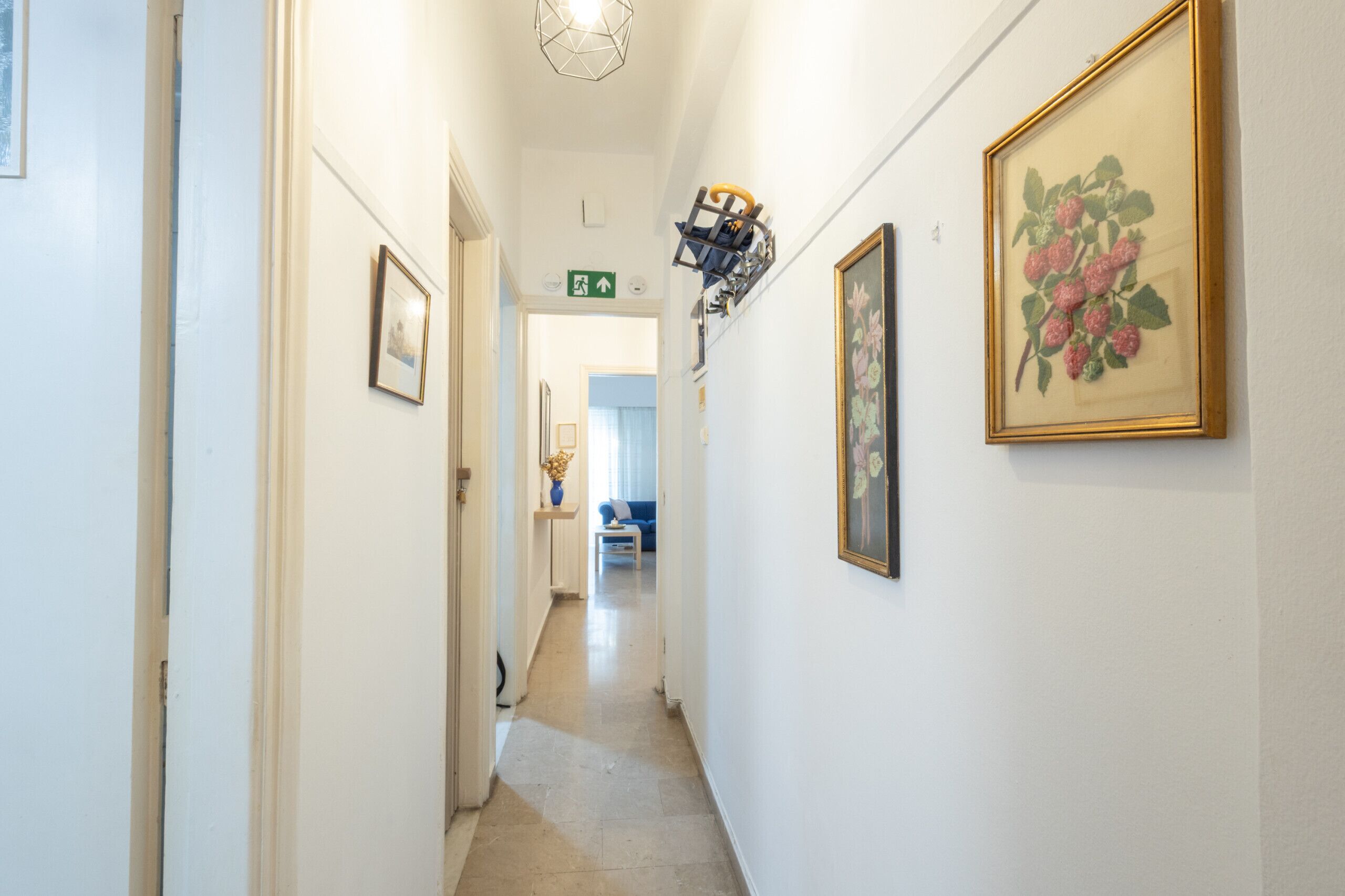A hallway with framed artwork, a green exit sign, and a blue couch in the background.
