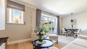 A modern living room with a round coffee table, a sofa, a dining table with chairs, and a large window with curtains. - Meersalz - Sea salt - Exclusive vacation apartment with sauna, terrace & fireplace in (Sylt)