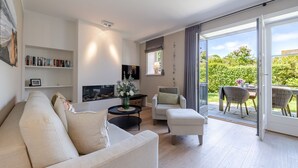 A modern living room with a sofa, armchairs, a coffee table, and a fireplace. There is a bookshelf and a TV mounted on the wall. The room has large sliding glass doors leading to an outdoor patio with a table and chairs. - Meersalz - Sea salt - Exclusive vacation apartment with sauna, terrace & fireplace in (Sylt)