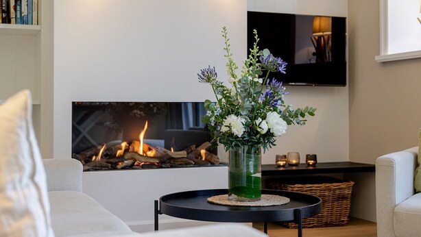 A living room with a fireplace, a flat-screen TV mounted on the wall, and a vase with flowers on a side table. - Meersalz - Sea salt - Exclusive vacation apartment with sauna, terrace & fireplace in (Sylt)