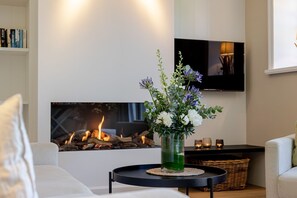 A living room with a fireplace, a flat-screen TV mounted on the wall, and a vase with flowers on a side table. - Meersalz - Sea salt - Exclusive vacation apartment with sauna, terrace & fireplace in (Sylt)