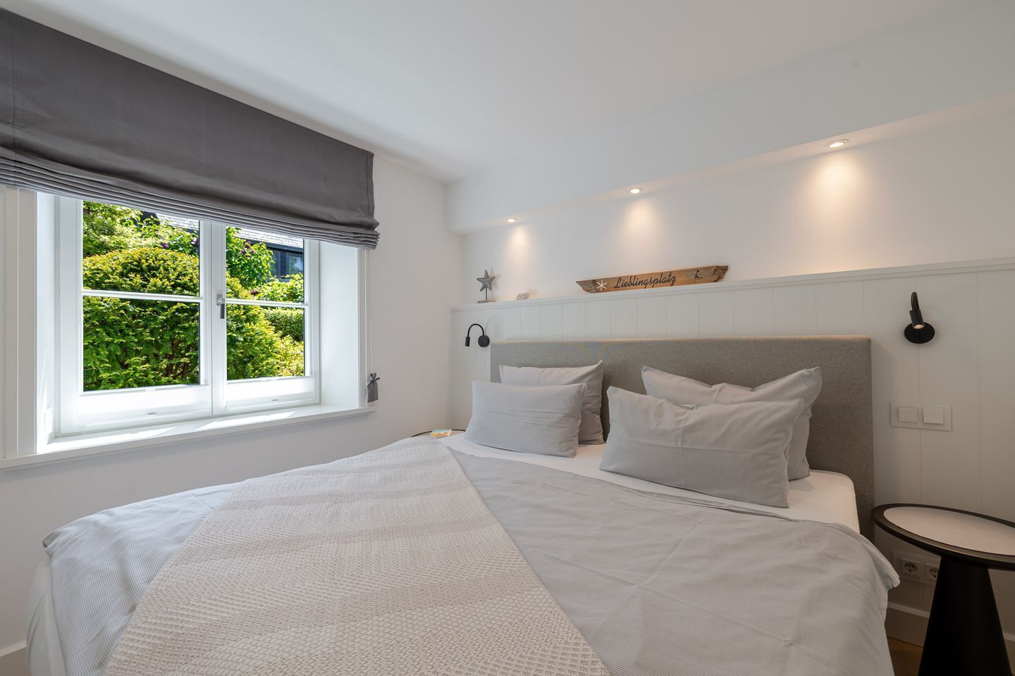 Une chambre avec un grand lit, une table de chevet, une fenêtre donnant sur un paysage verdoyant et un luminaire fixé au mur.