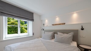 A bedroom with a large bed, a bedside table, a window with a view of greenery, and a wall-mounted light fixture. - Meersalz - Sea salt - Exclusive vacation apartment with sauna, terrace & fireplace in (Sylt)