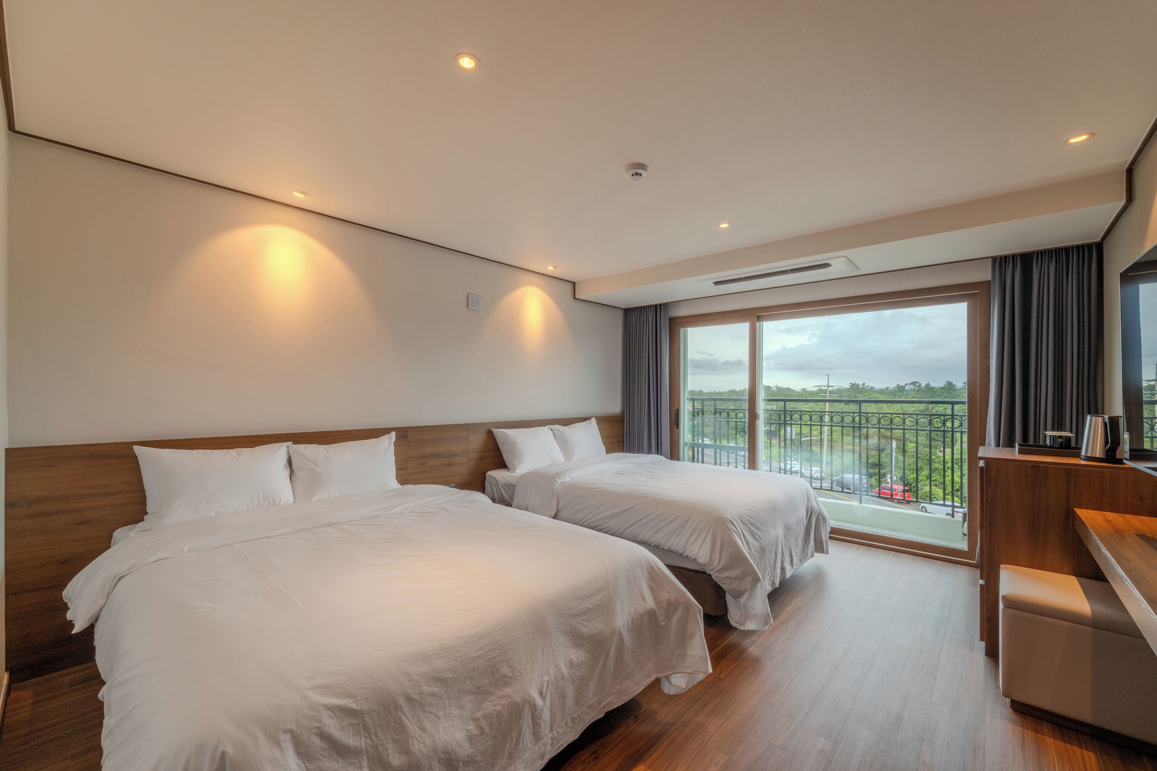 A hotel room with two beds, a wooden headboard, a large window with a balcony, and a desk with a chair.