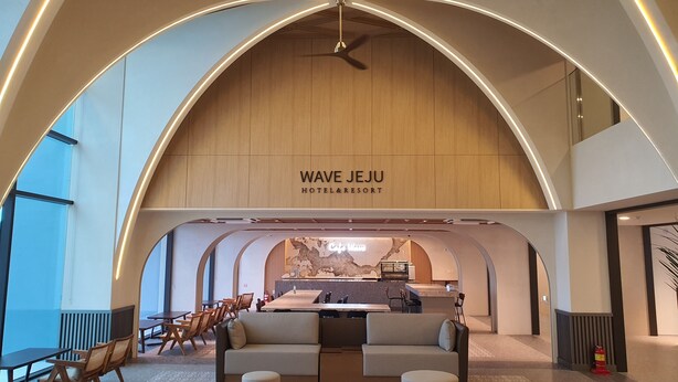 A modern hotel lobby with a high ceiling, a central seating area, and a visible kitchen in the background.