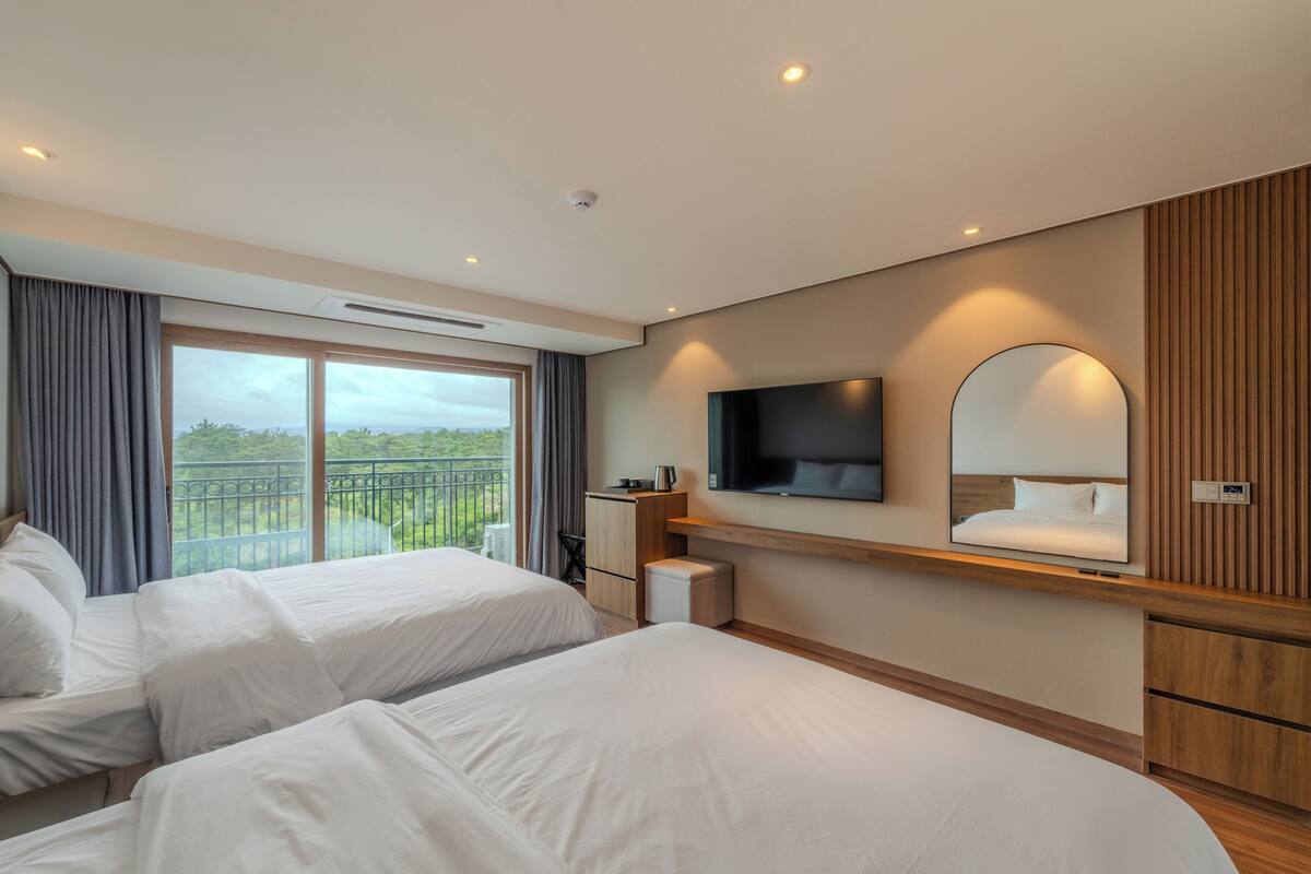 Habitación de hotel con dos camas, televisión, espejo y balcón con vistas a zonas verdes.