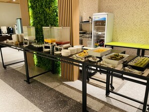 Montaje de bufé con diferentes alimentos y bebidas, que incluye un dispensador de jugo y un refrigerador con la etiqueta 'UNICOLA'.