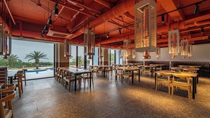 A spacious restaurant with high ceilings, exposed pipes, and large windows overlooking a pool.